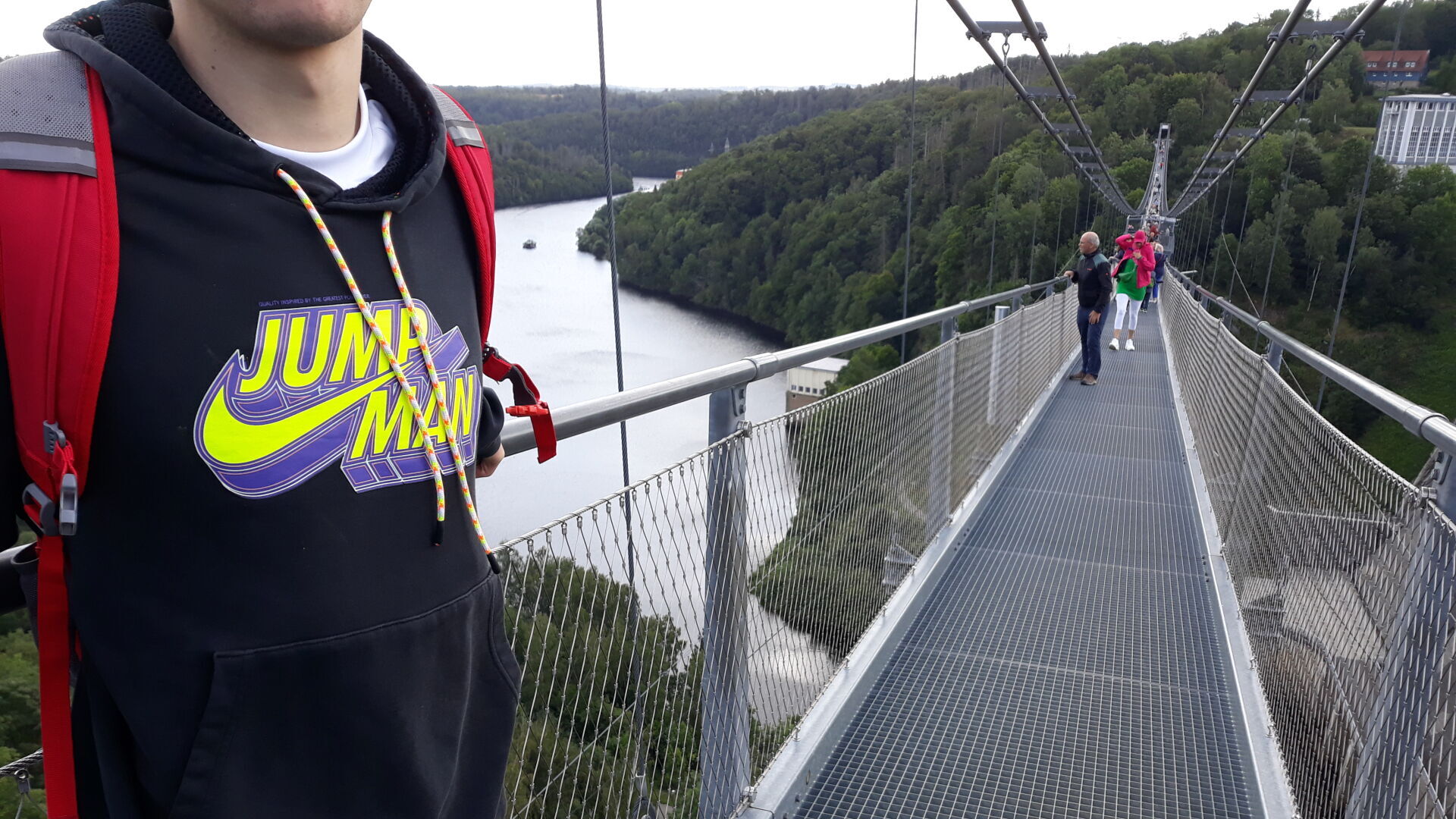 Auf langen Hängebrücke über Flusstal steht junger Mann mit Nike-Pullover "Jump Man ✓"